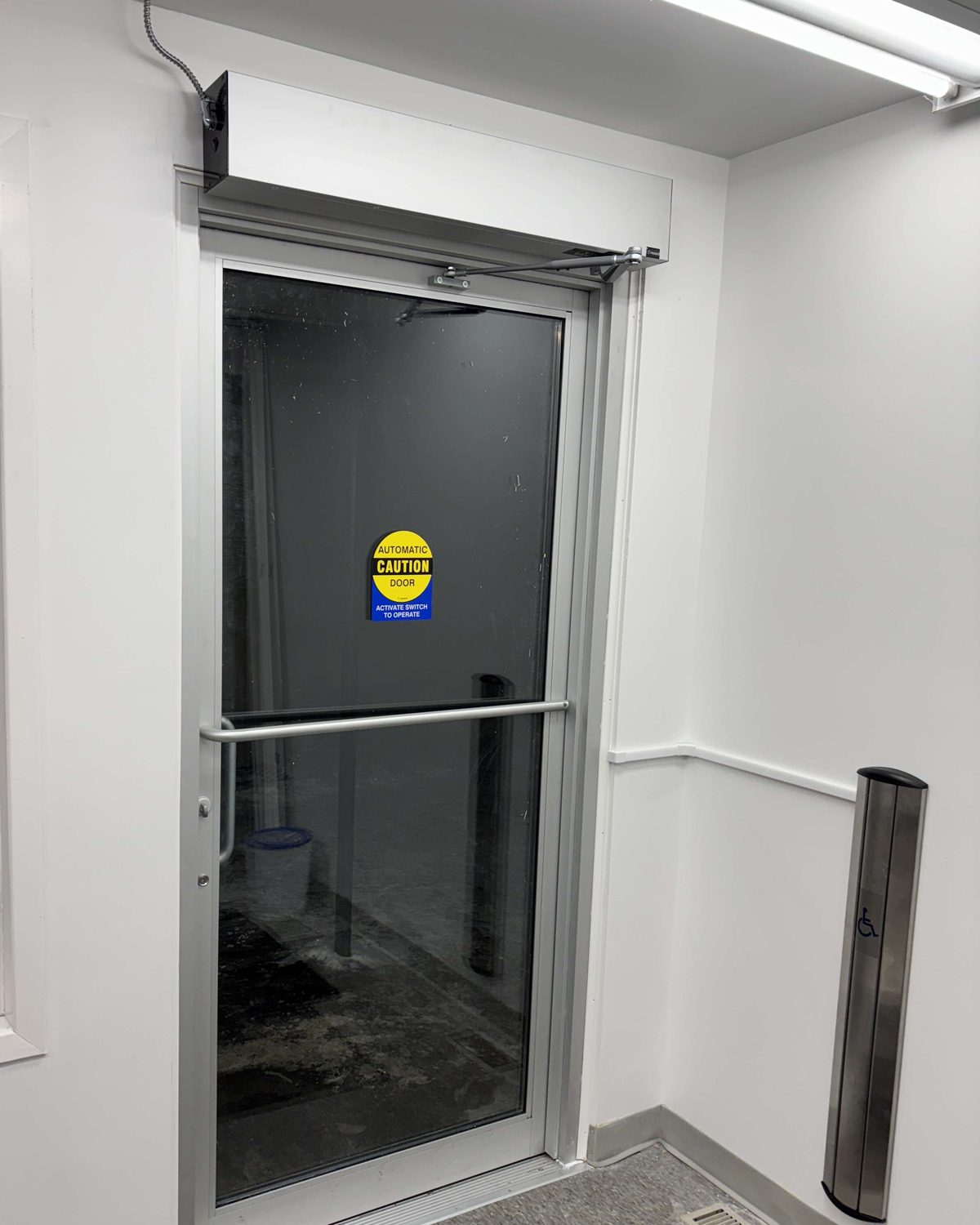 A clear aluminum glass door with a horizontal push bar, an automatic operator in a white header box above it, an automatic door caution decal on the glass and a stainless push-button column on the wall beside it