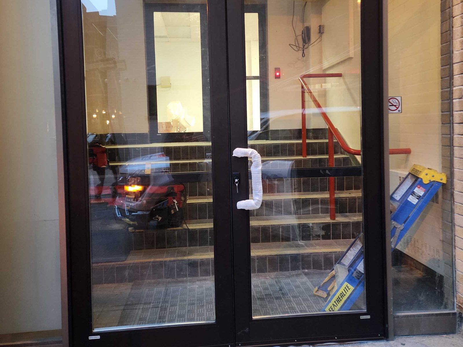 A new pair of dark-finished aluminum storefront doors at the Omni King Edward Hotel in Toronto, the centre pull still wrapped in protective foam, with fixed glass either side and an interior stair visible through the glass