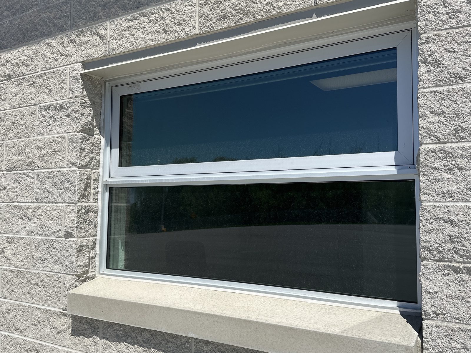 A white-framed aluminum commercial window set into a split-face concrete block wall above a precast sill, seen at an angle, with an operable vent over a large fixed lite