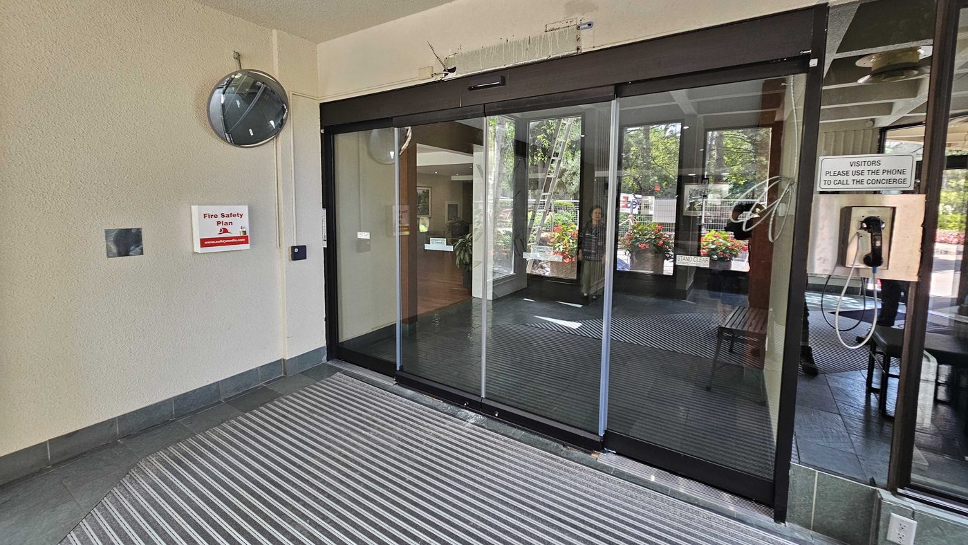 An all-glass automatic sliding door in dark aluminum framing at a condominium lobby entrance, with a wide recessed metal grille entrance mat directly in front of the leaves and the operator header above them