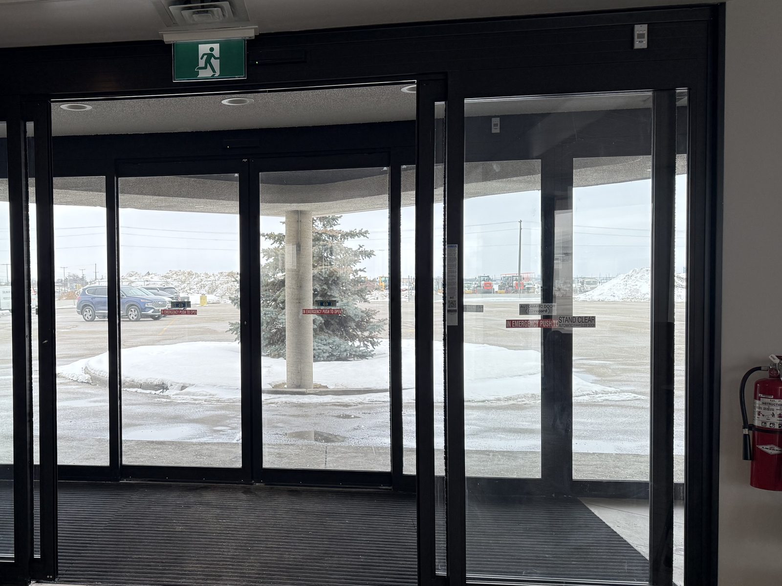 A finished glass vestibule seen from inside, with two sets of automatic sliding doors in black aluminum framing under an exit sign, Record automatic-door and emergency push-to-open decals on the glass, a recessed entrance mat between them and a snowy parking lot beyond