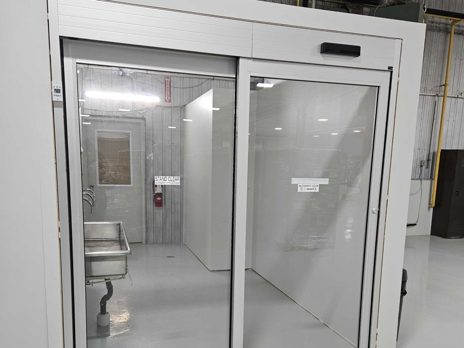 A single-slide automatic door in white aluminum framing inside an industrial plant, with a black sensor mounted on the right-hand end of the header and a STAND CLEAR decal on the glass