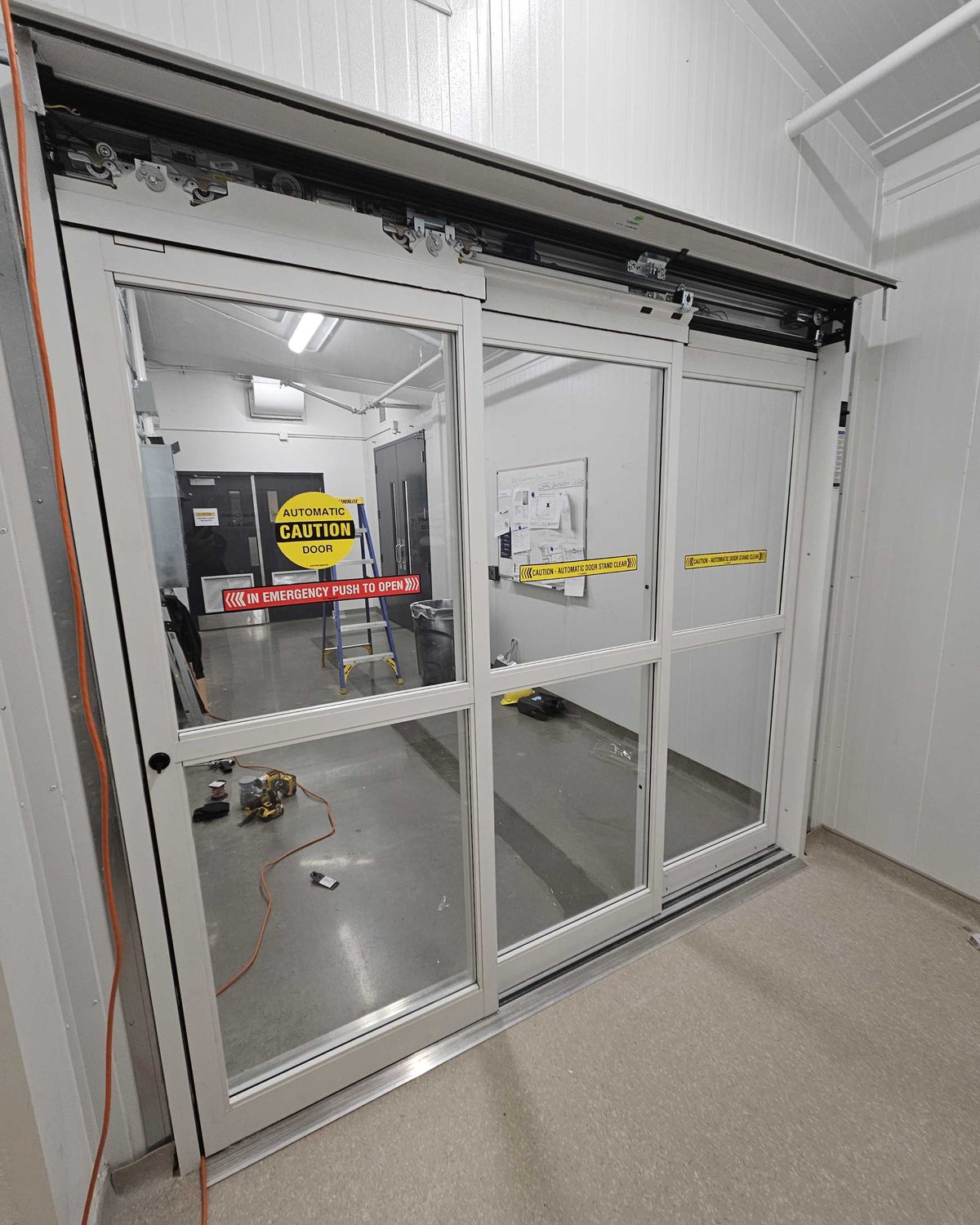 A three-panel automatic sliding door in white aluminum framing during installation, the header cover off to show the track and the carriers the leaves hang from, with automatic-door caution and emergency push-to-open decals on the glass and tools on the floor