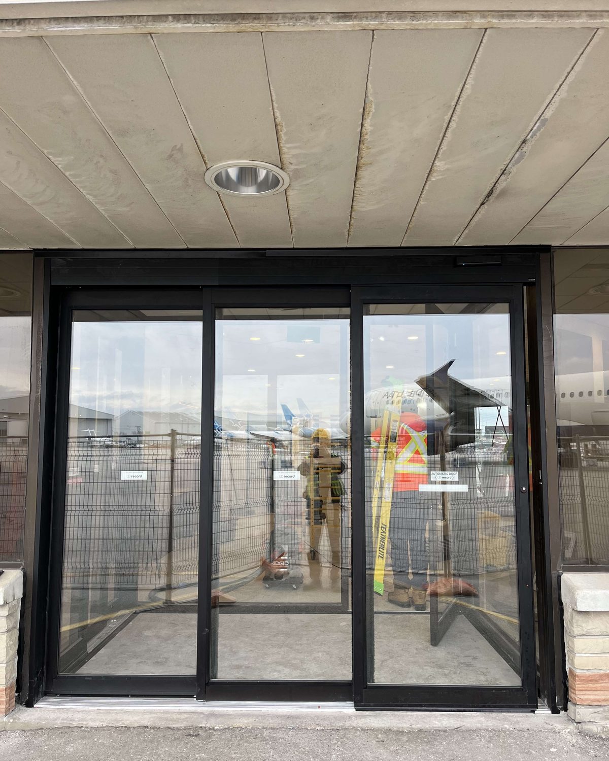 A three-panel automatic sliding entrance in black aluminum framing under a concrete canopy at an aviation facility in Mississauga, with construction fencing and caution tape still visible through the glass