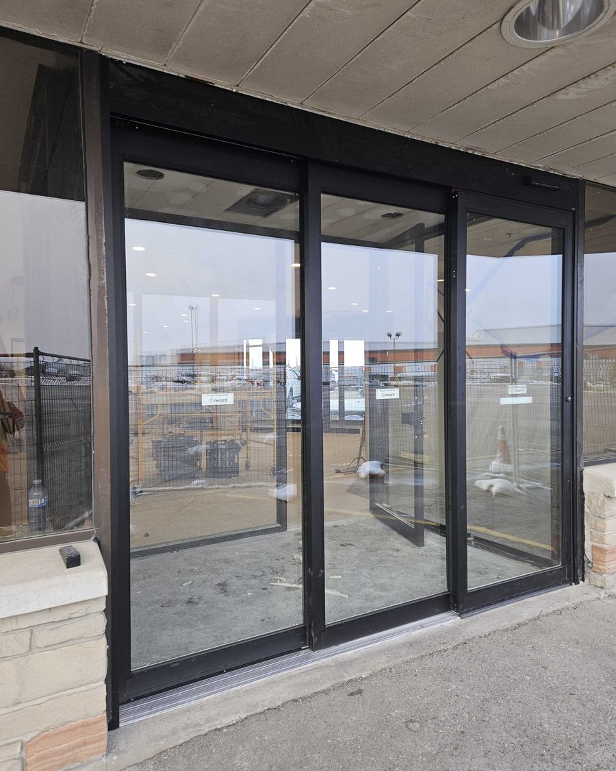 A newly installed three-panel automatic sliding door in black aluminum framing with Record decals on the glass, under a concrete canopy at an aviation facility in Mississauga, the vestibule behind it still under construction