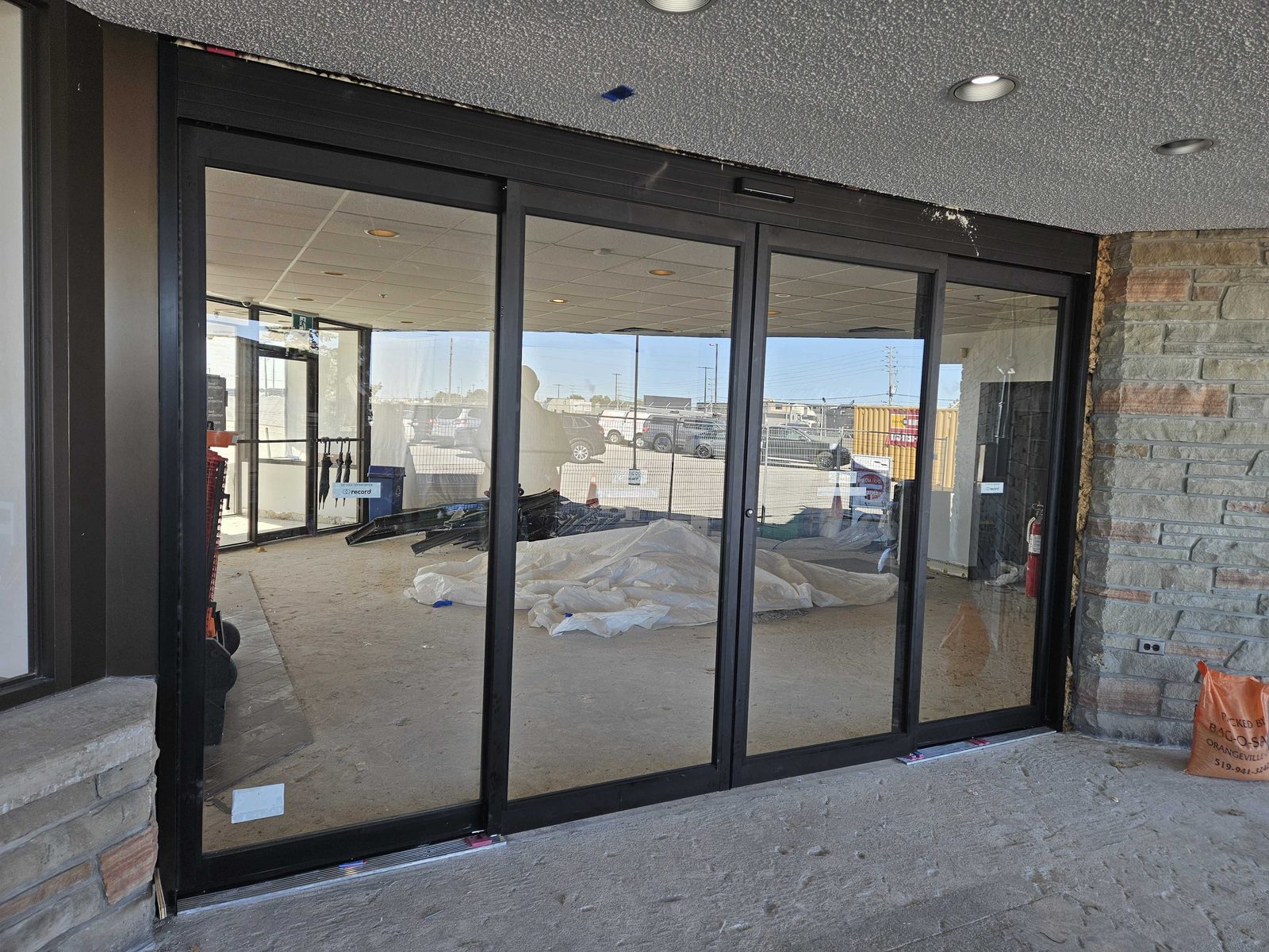 A new four-leaf automatic sliding entrance in dark aluminum framing being installed under a textured canopy soffit between stone-clad piers, the floor inside still unfinished and covered with protective sheeting