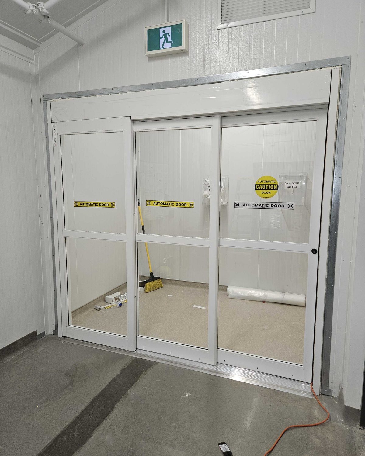 A white-framed automatic sliding door with three glazed panels inside an industrial building, AUTOMATIC DOOR decals across the leaves and a yellow caution sticker, an exit sign above the header