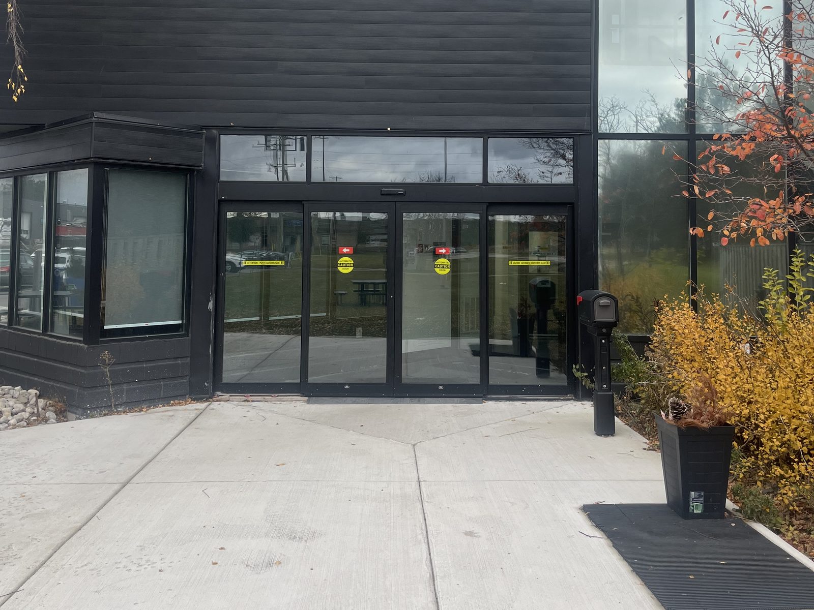 A black anodized automatic sliding entrance in a dark-clad building, its two centre leaves carrying yellow caution decals between fixed glass side panels, with a concrete walkway and autumn shrubs in front