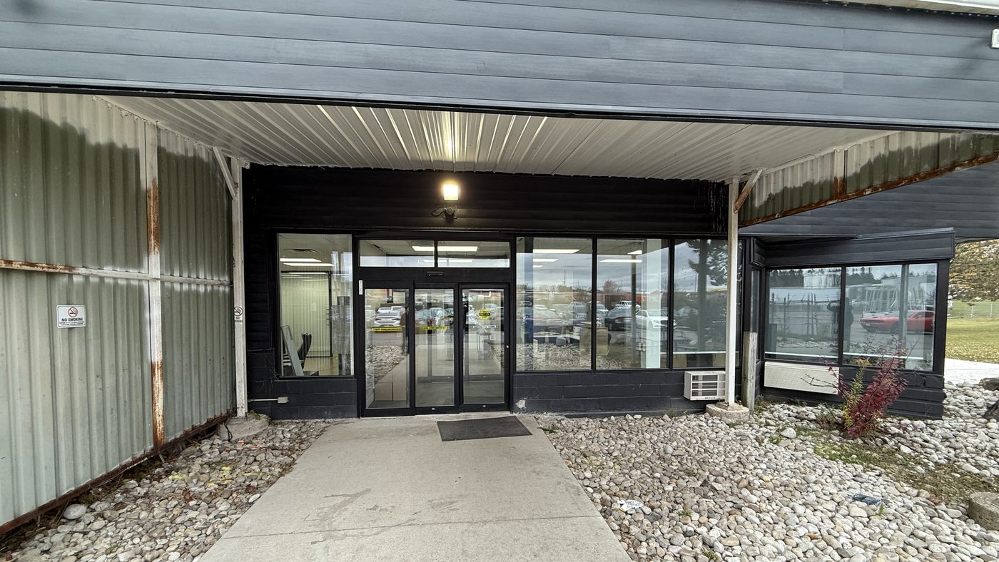 The entrance of a dark-clad industrial building under a metal canopy, with an automatic sliding door in black framing set between wide glazed panels at the end of a concrete walkway lined with river stone