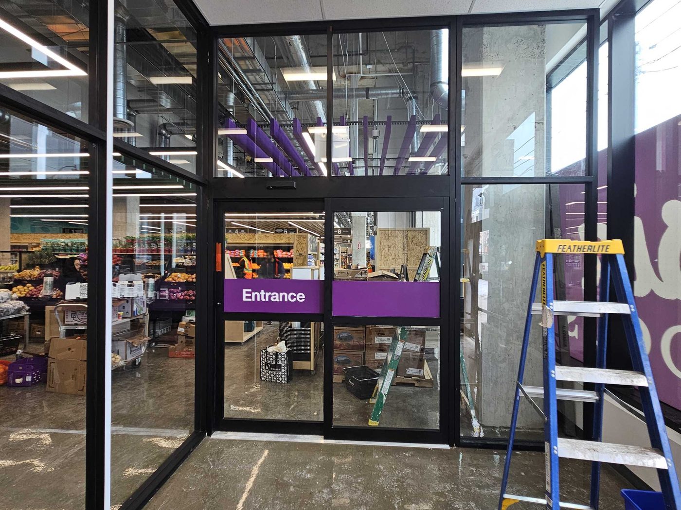 A black aluminum storefront vestibule inside a grocery store with an automatic sliding entrance marked Entrance, fixed glass above and to the side, and a step ladder standing on the new floor