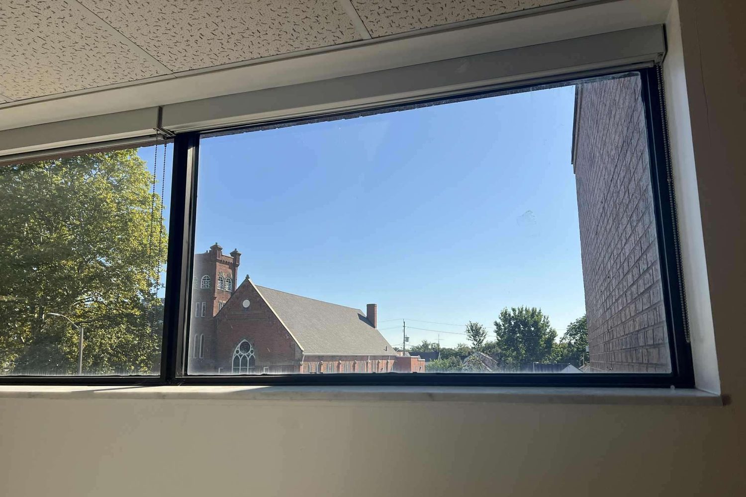 Seen from inside an office, a long run of black-framed fixed aluminum windows under a suspended ceiling, looking out to a brick church and trees