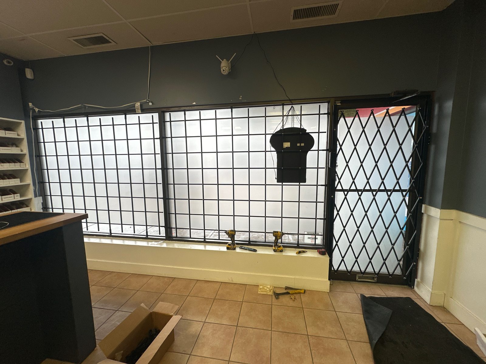 Inside a retail unit, black steel bars fixed over a run of frosted storefront windows and a black expanding diamond-lattice gate across the glass entrance door, with cordless drills still on the window sill and a security camera on the wall above