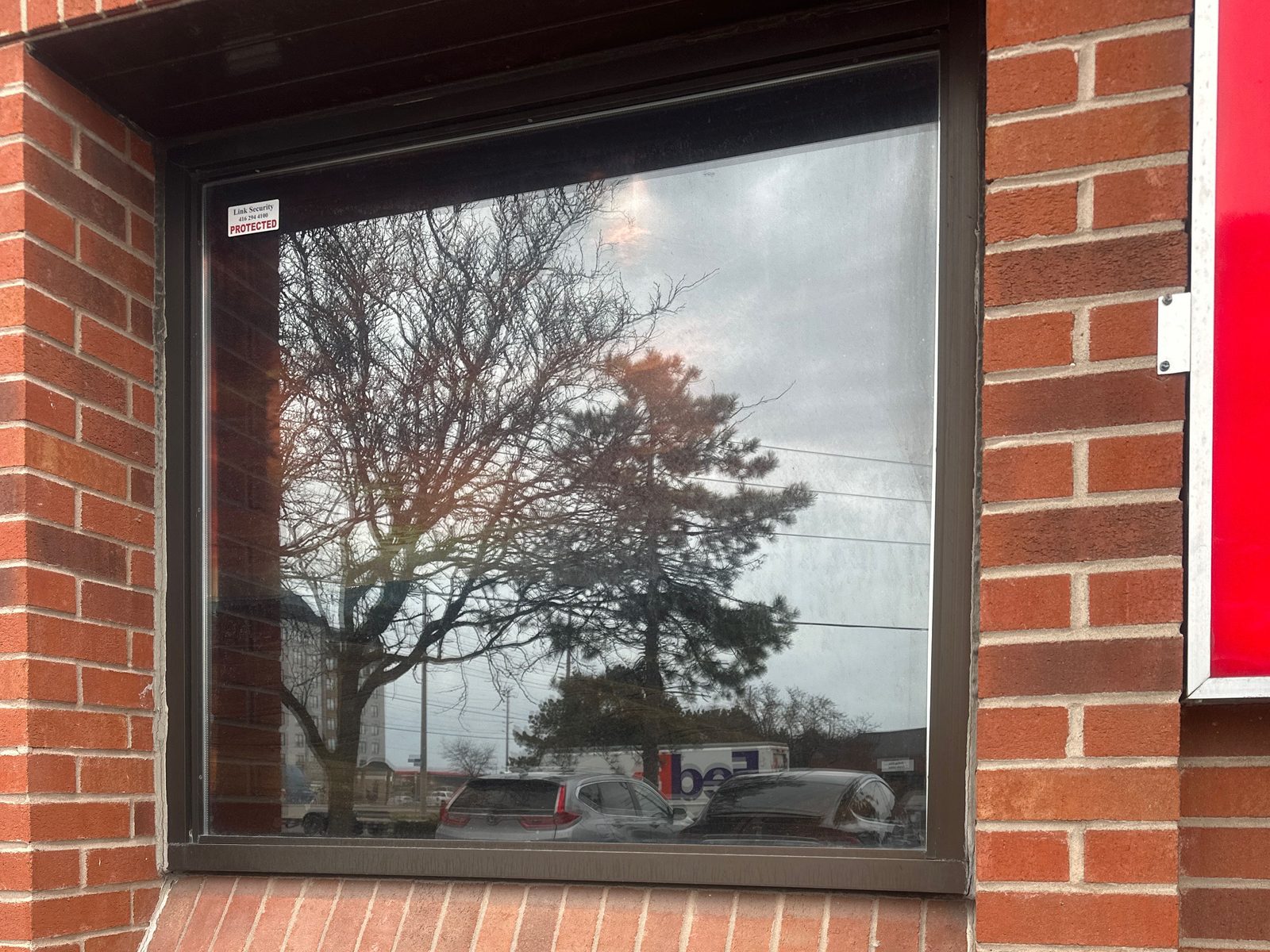A single large fixed window in a dark bronze aluminum frame set into a red brick wall of a commercial building, a security company decal in the corner of the glass and trees and parked cars reflected in it