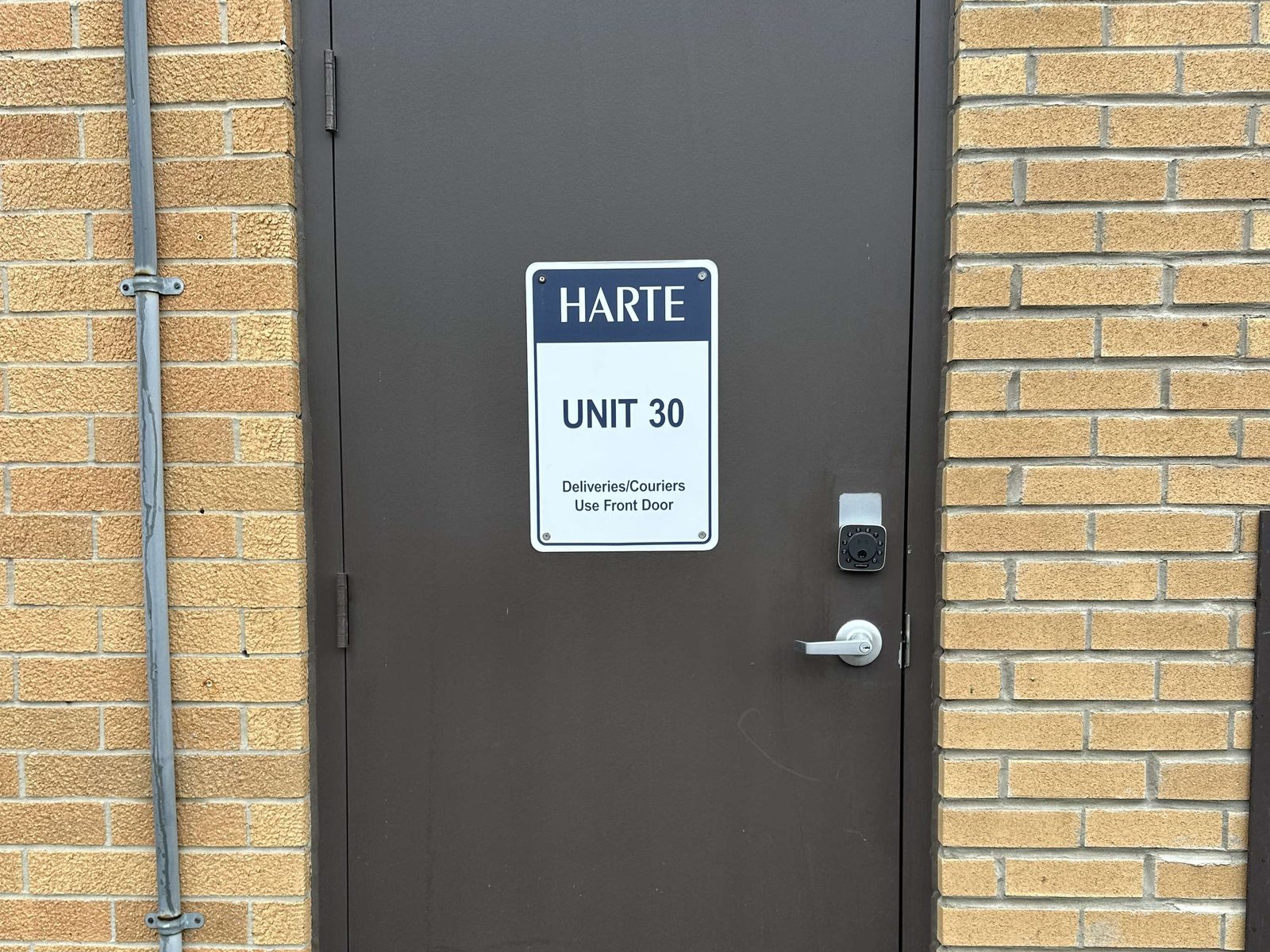 A dark brown steel door in a yellow brick wall, fitted with a keypad lockset above a lever handle beside a tenant sign reading Unit 30, with a conduit running up the wall beside the frame