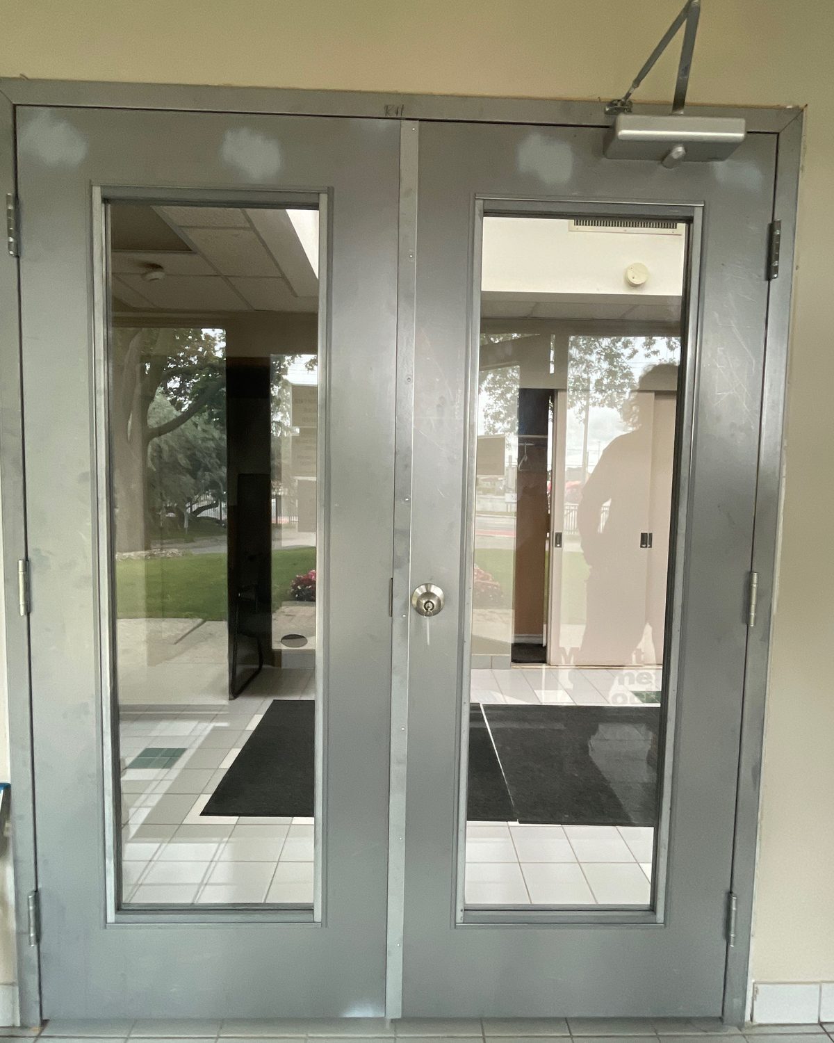 A pair of grey steel doors with full-height glass lites and a surface closer, seen from outside looking into a tiled entrance lobby