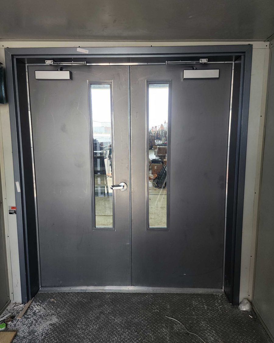 A pair of dark grey steel doors with narrow vision lites and a surface closer on each leaf, set in a wide steel frame