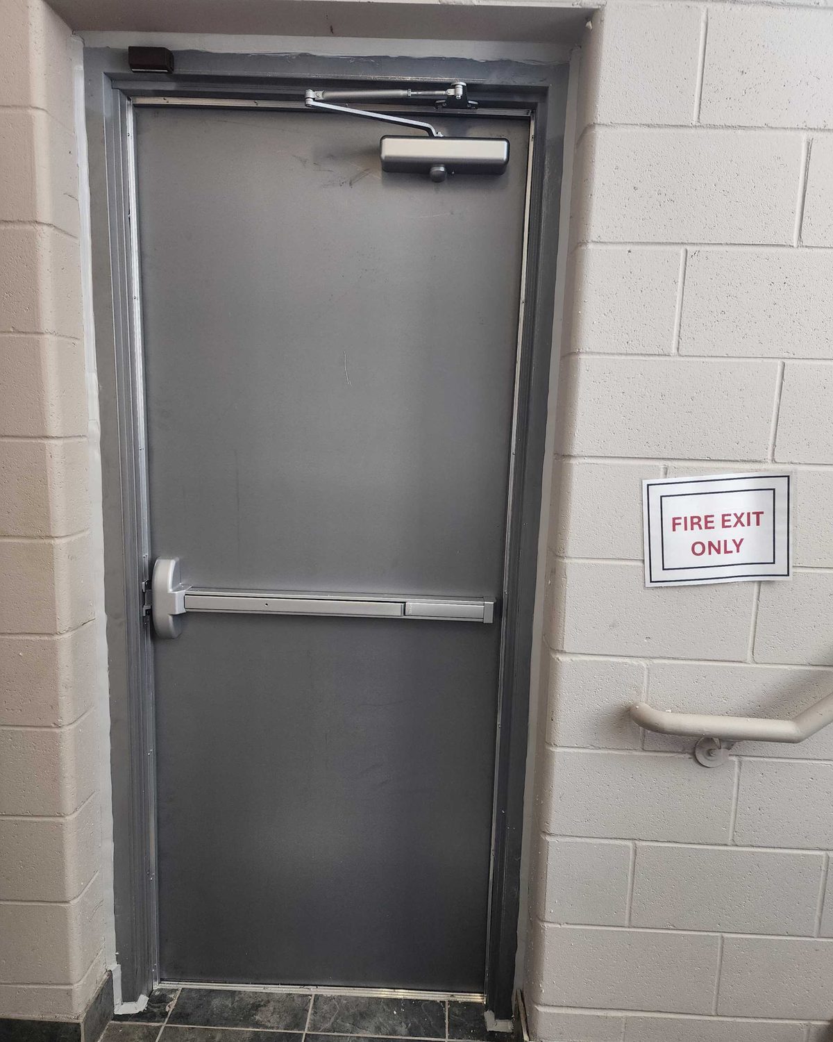A grey steel exit door with a horizontal push bar and a surface closer, set in a painted block wall beside a sign reading Fire Exit Only