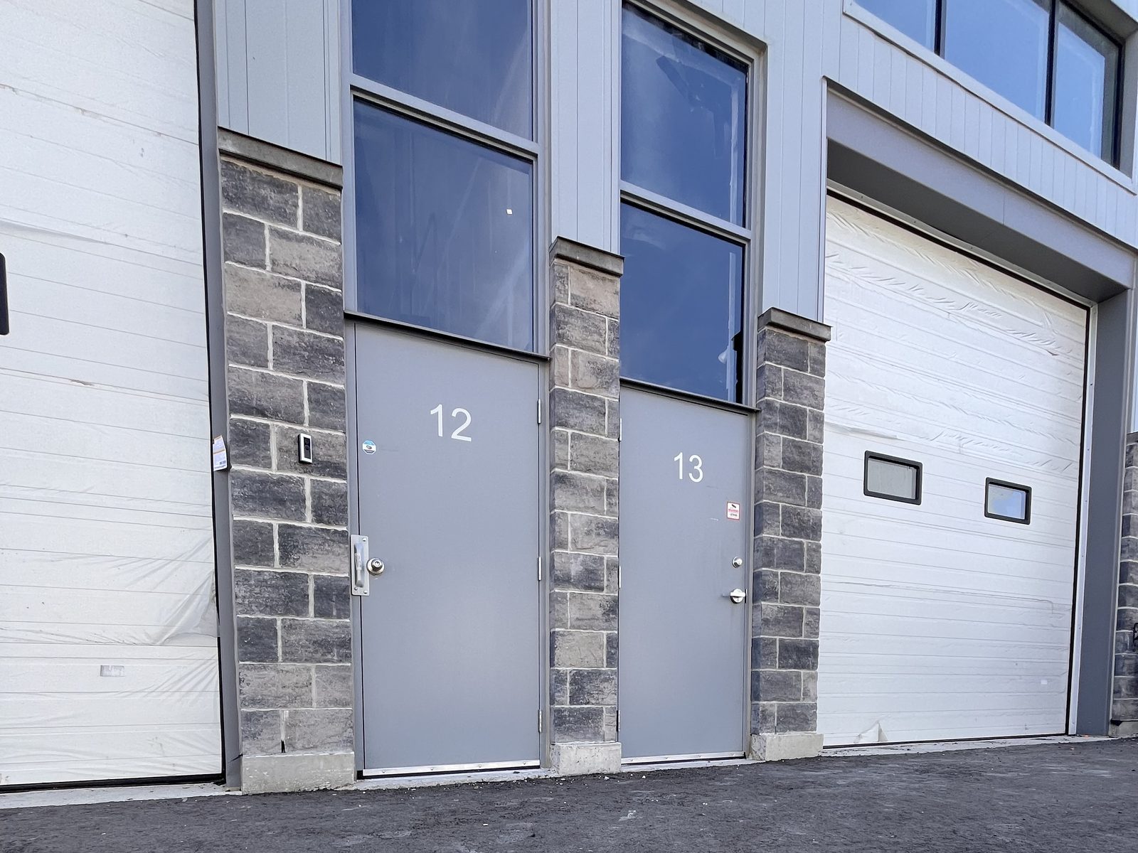 Two grey steel pedestrian doors numbered 12 and 13 set into a stone-clad industrial unit wall between two white sectional overhead doors, with tall windows above each steel door
