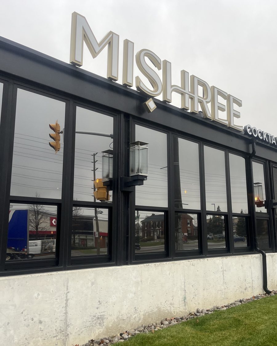 The glazed corner of the Mishree restaurant in Mississauga seen from below, black aluminum mullions running the full height of the glass beneath the illuminated sign on the fascia