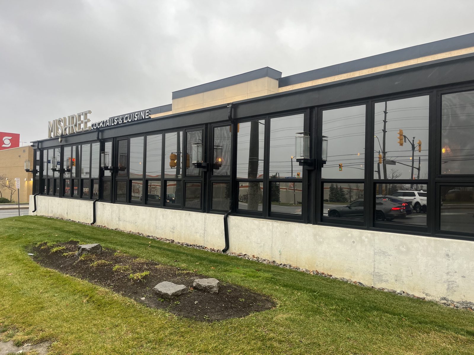 A single-storey restaurant glazed along its full length in black aluminum framing — tall fixed lites over a row of smaller ones, wall lanterns mounted between the mullions — sitting above a concrete kerb wall, with the MISHREE Cocktails & Cuisine sign along the fascia