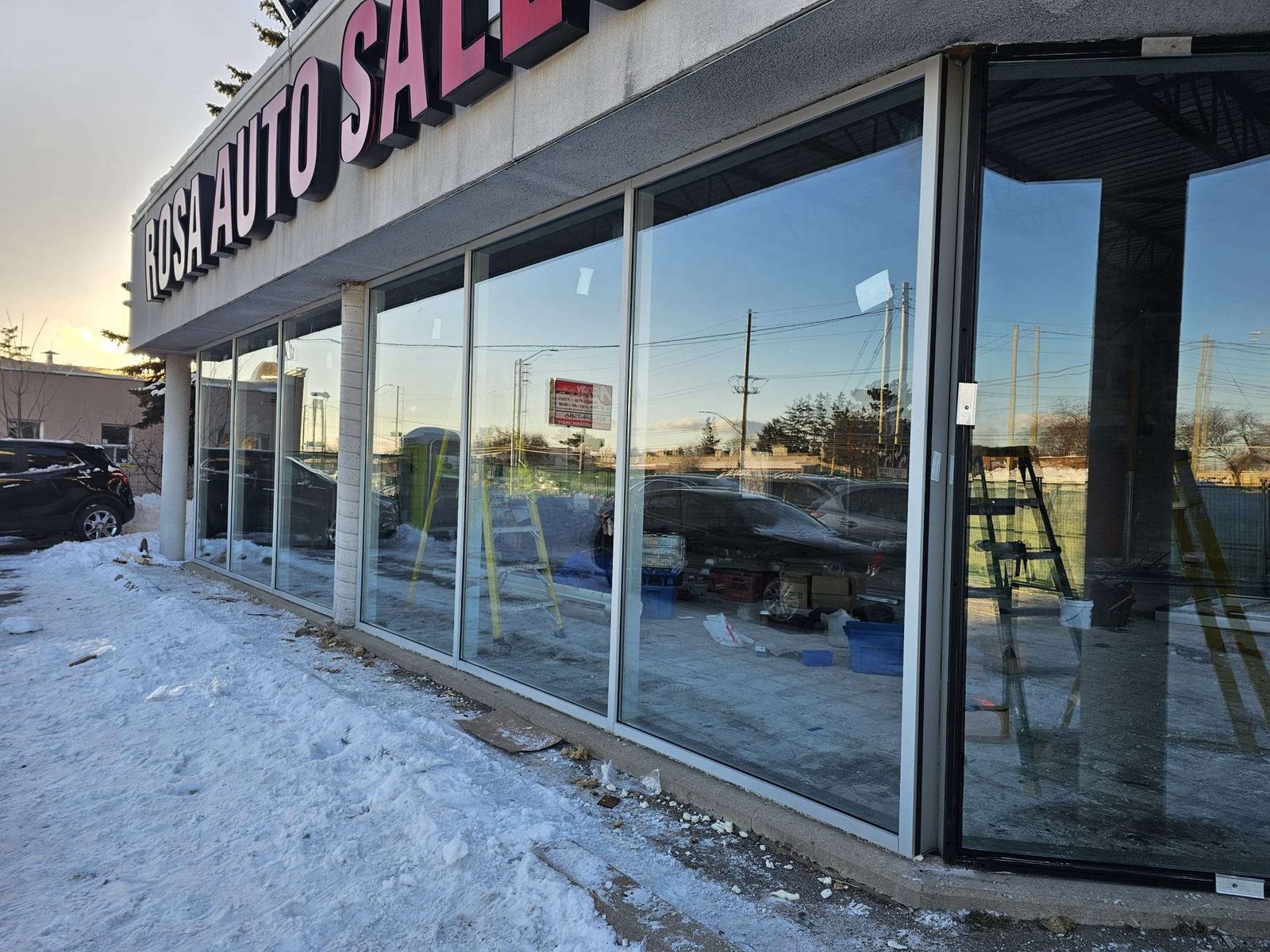 A run of newly installed white-framed aluminum storefront glazing along a car dealership in winter, protective film still on the glass and step ladders standing inside the empty showroom