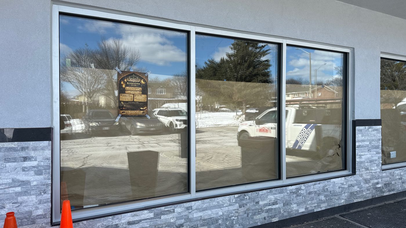 A large three-lite fixed commercial window assembly in clear anodized aluminum framing above a grey stacked-stone base, protective film still on the glass, with a Canuck Door Systems service truck reflected in it