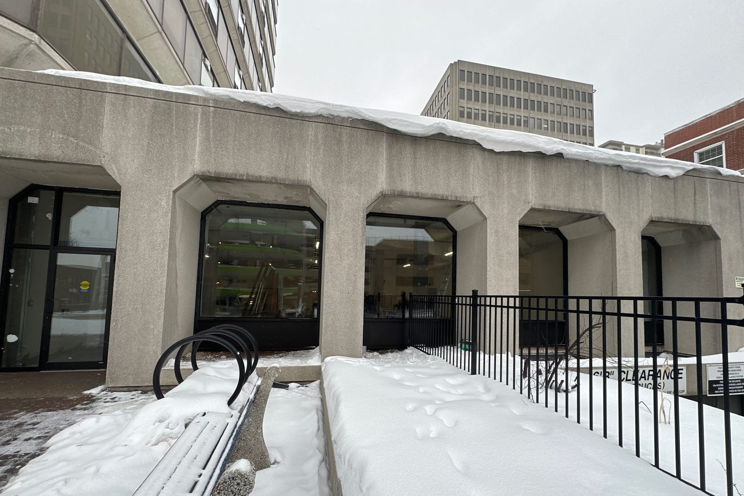 Black-framed commercial window units set into the chamfered concrete openings of a downtown office building in winter, snow banked along the wall and an interior fit-out visible through the glass