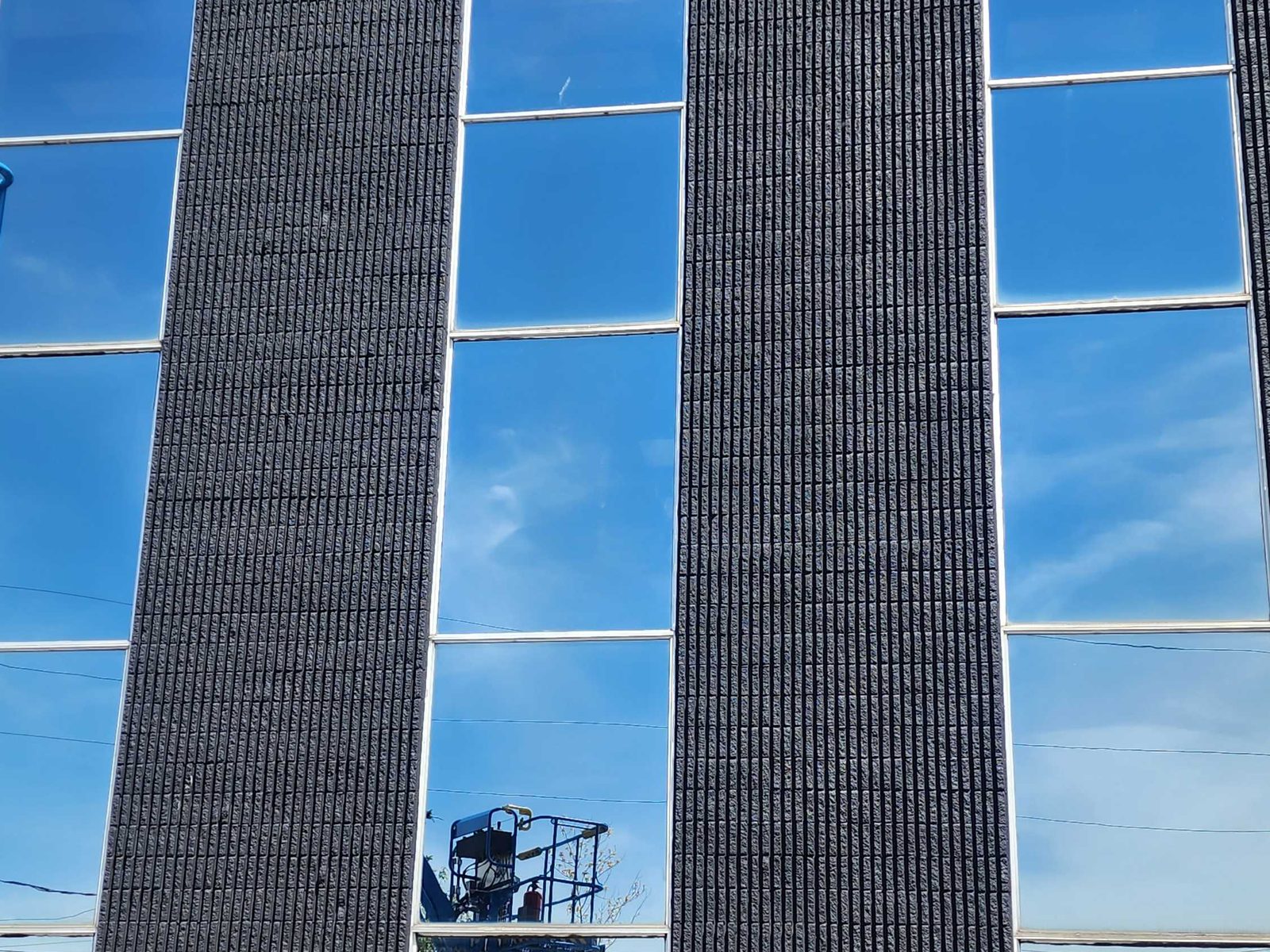 A multi-storey commercial elevation where tall bands of blue-reflective glass in white aluminum framing run unbroken past the floor lines between dark split-face block piers, with an aerial work platform reflected in the glass
