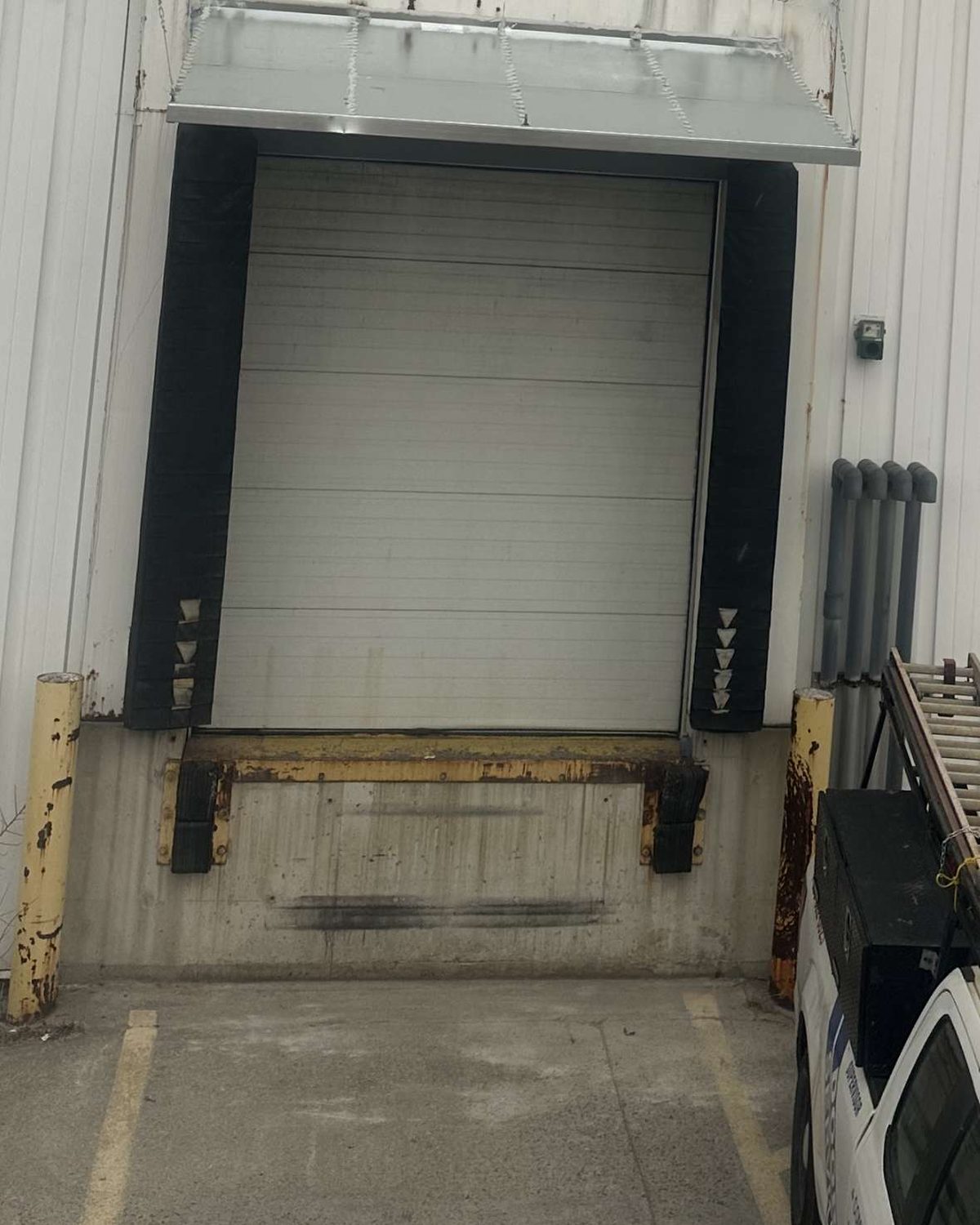 A dock position in a white metal-clad wall under a steel canopy, with a white sectional door between black shelter side pads, a worn yellow leveler frame at the sill and yellow bollards either side