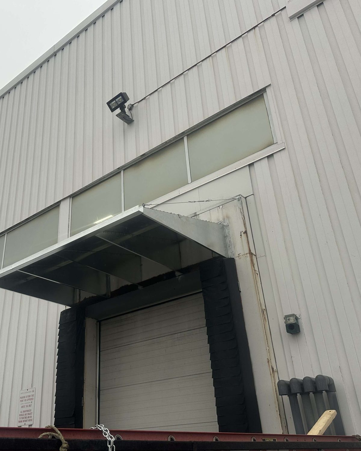 Looking up at a dock position on a white metal-clad warehouse: a steel canopy projecting over a black dock shelter and a white sectional door, with clerestory windows above