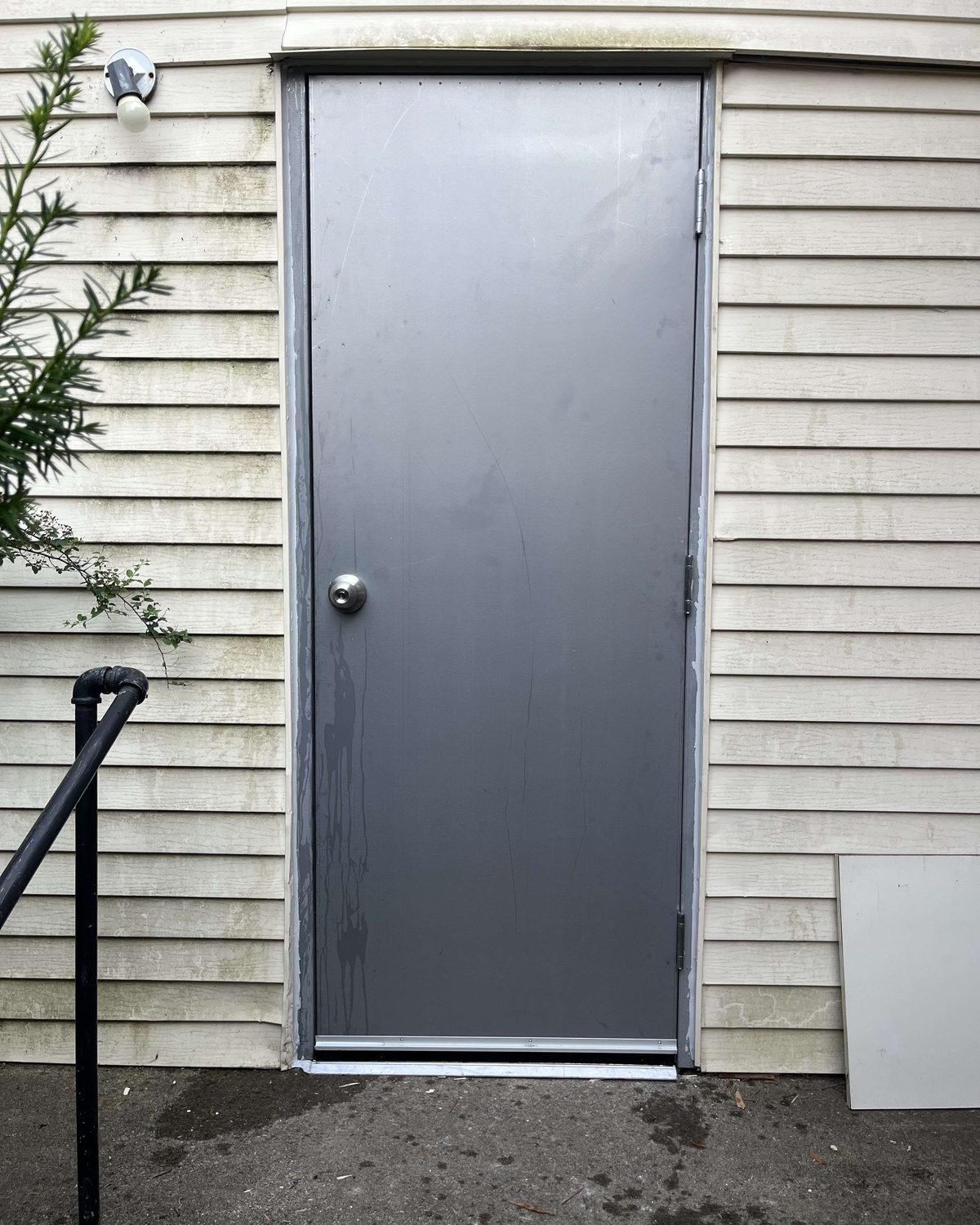 A plain flush grey steel door with a knob set, installed in a small building clad in cream vinyl siding