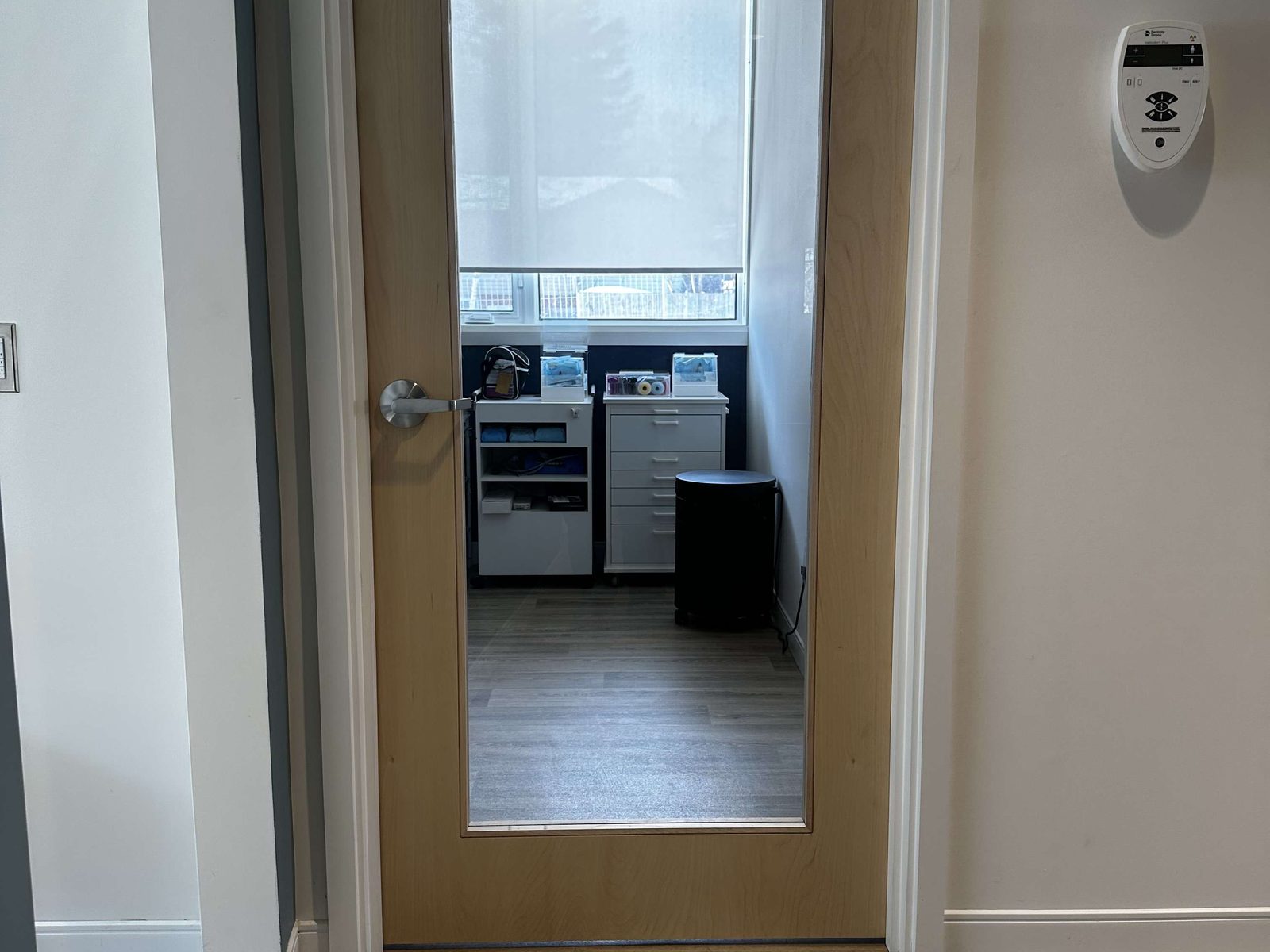 A flush light wood-veneer door with a large clear glass lite and a lever handle, set in white trim above a plank floor, with a small clinic room and a window blind visible through the glass