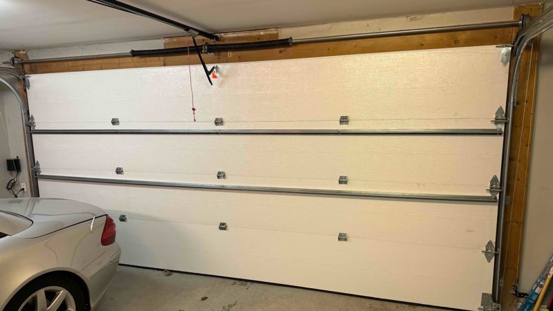 A white insulated double garage door seen from inside a garage, with horizontal reinforcing struts across its sections, the torsion spring on its shaft above the door head, the opener rail and red release cord, and a silver car parked alongside
