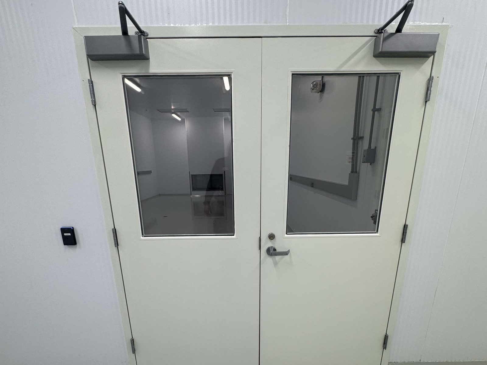 A pair of white hollow metal doors with large glass vision lites, two surface closers on the top rail and a lever handle, set in a white-panelled corridor with a small black access reader on the wall beside them