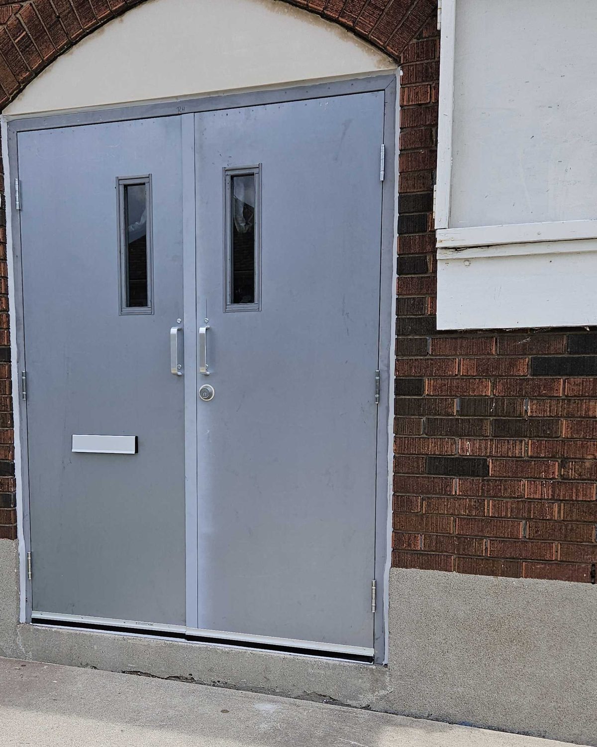 A pair of grey exterior hollow metal doors with narrow vision lites, pulls and a mail slot, set in a brick wall under a rendered arch