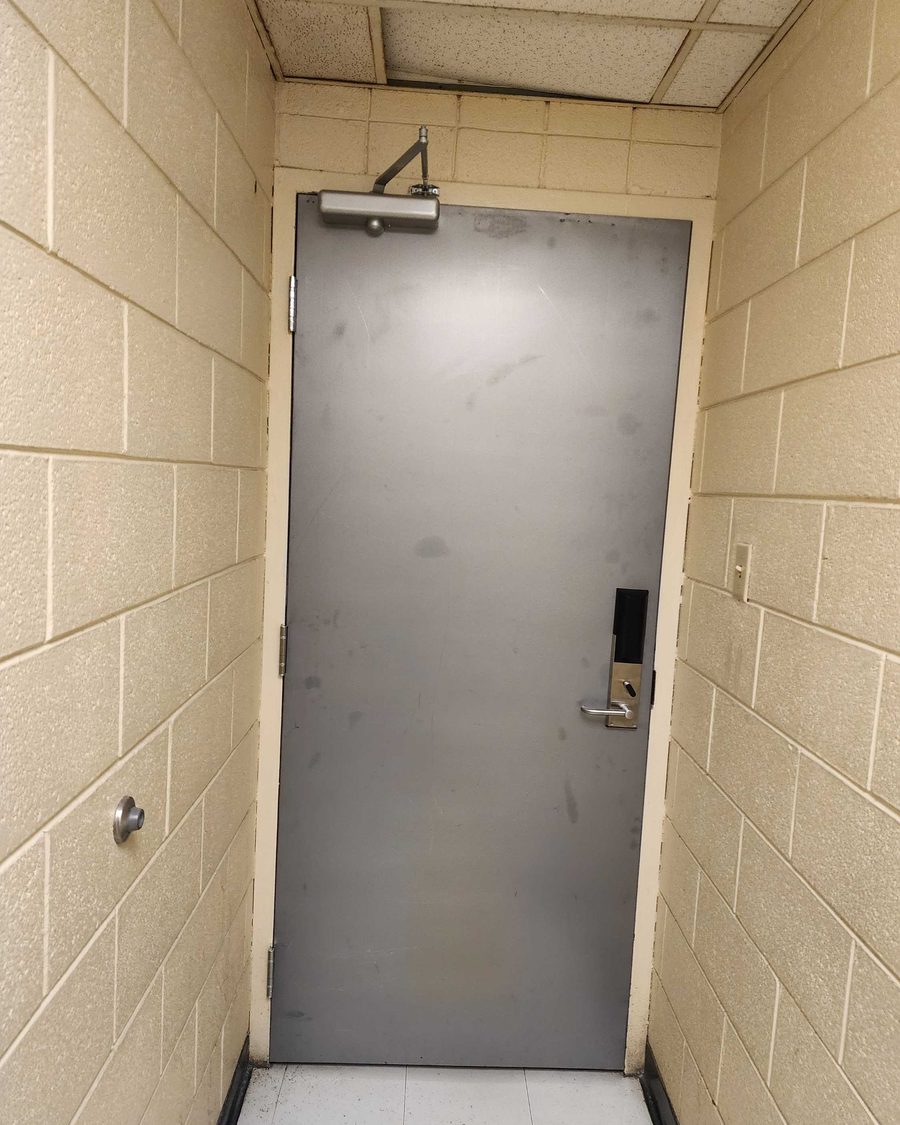 A grey steel door at the end of a painted block-walled hotel service corridor, carrying an electronic card-reader lockset and a surface-mounted closer at the head