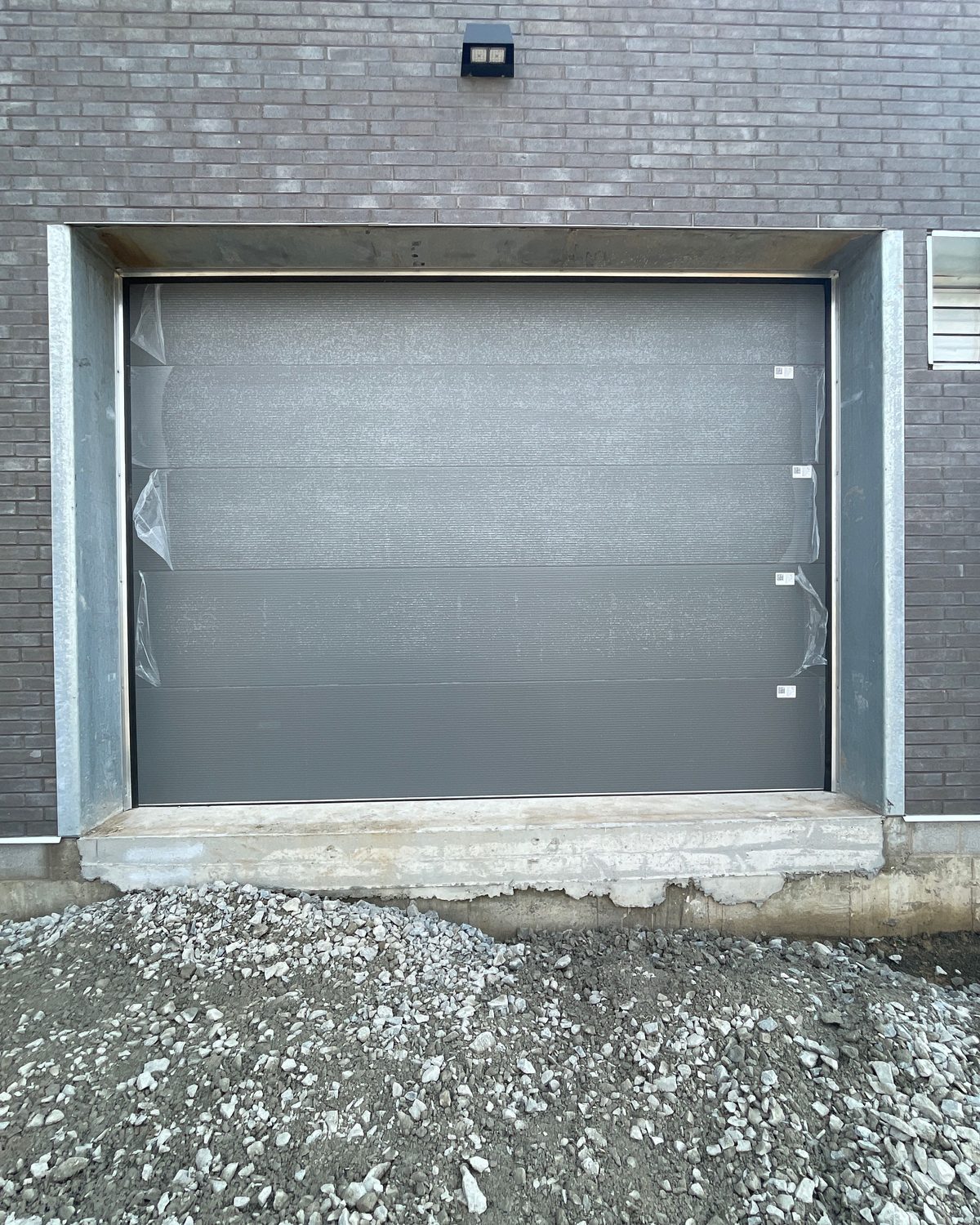 A grey insulated sectional overhead door with protective film still on its panels, set in a galvanized steel frame in a new dark brick wall above a raised concrete sill