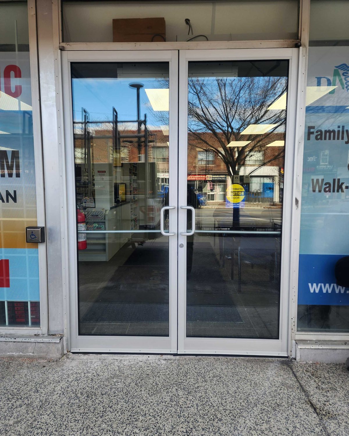 A pair of clear anodized narrow-stile aluminum storefront doors with offset pulls at a medical clinic, a wall-mounted accessibility push button beside the frame and an automatic door caution decal on the glass