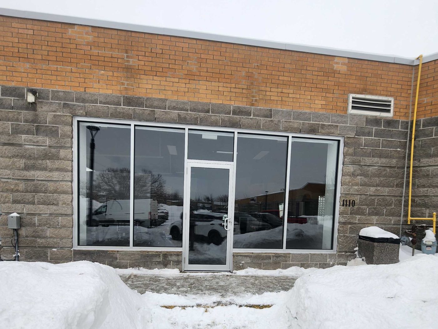 A new aluminum storefront — fixed glass lites either side of a single glass door — on a block and brick commercial unit in winter, with a path cut through deep snow banks to the entrance