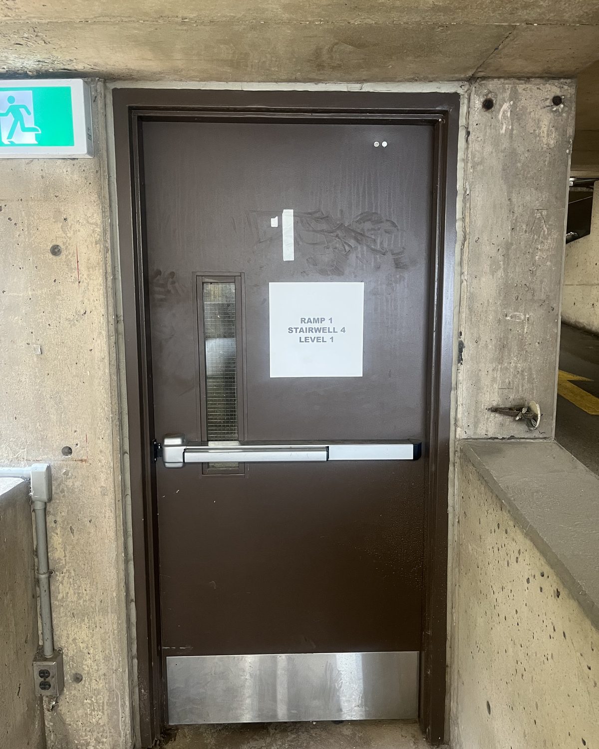 A brown-painted hollow metal stairwell door with a narrow vision lite, a panic bar and a stainless kick plate, in a concrete parking garage wall beside an exit sign