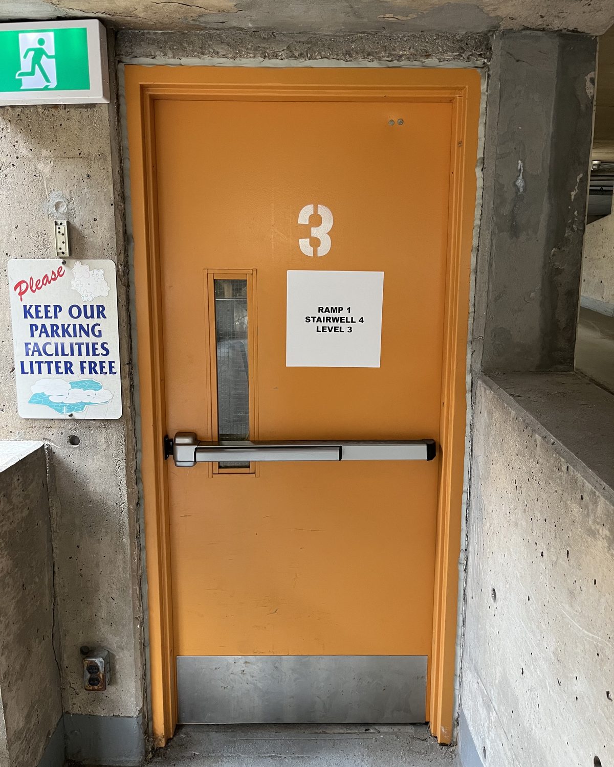 An orange-painted hollow metal stairwell door with a narrow vision lite, a panic bar, a stainless kick plate and a large stencilled number 3, set in a concrete parking garage wall