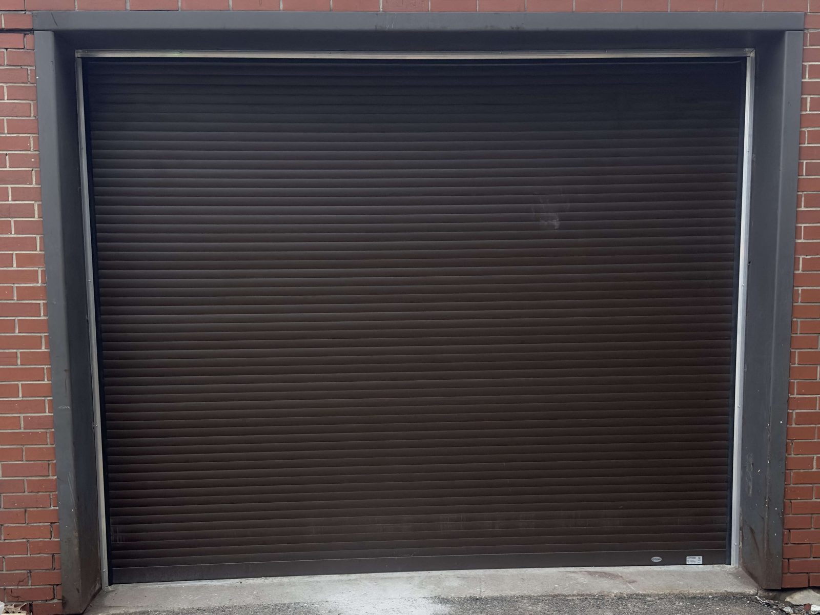 A dark brown roll-up door closed in a dark grey steel-framed opening in a red brick wall, its slat curtain running down to a concrete sill