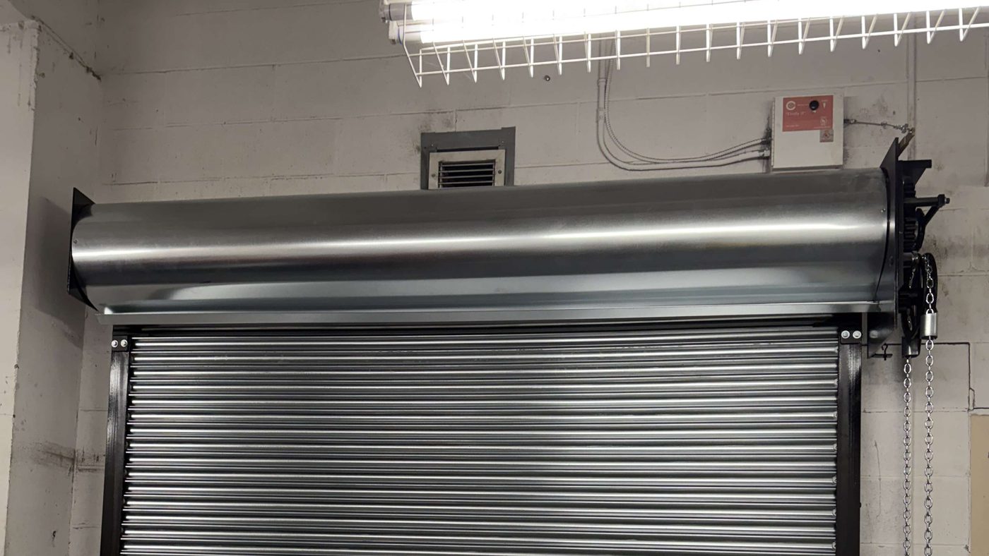 The head of a galvanized rolling door inside a painted block wall: a curved steel hood over the coiled curtain, the curtain running down into its guides, and a hand chain hanging at the right-hand side