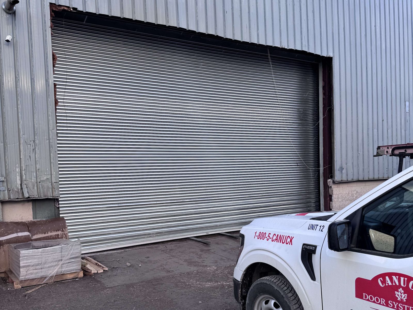 A wide galvanized rolling steel door closed in the corrugated metal wall of an industrial unit, with a white Canuck Door Systems service pickup parked in front of it