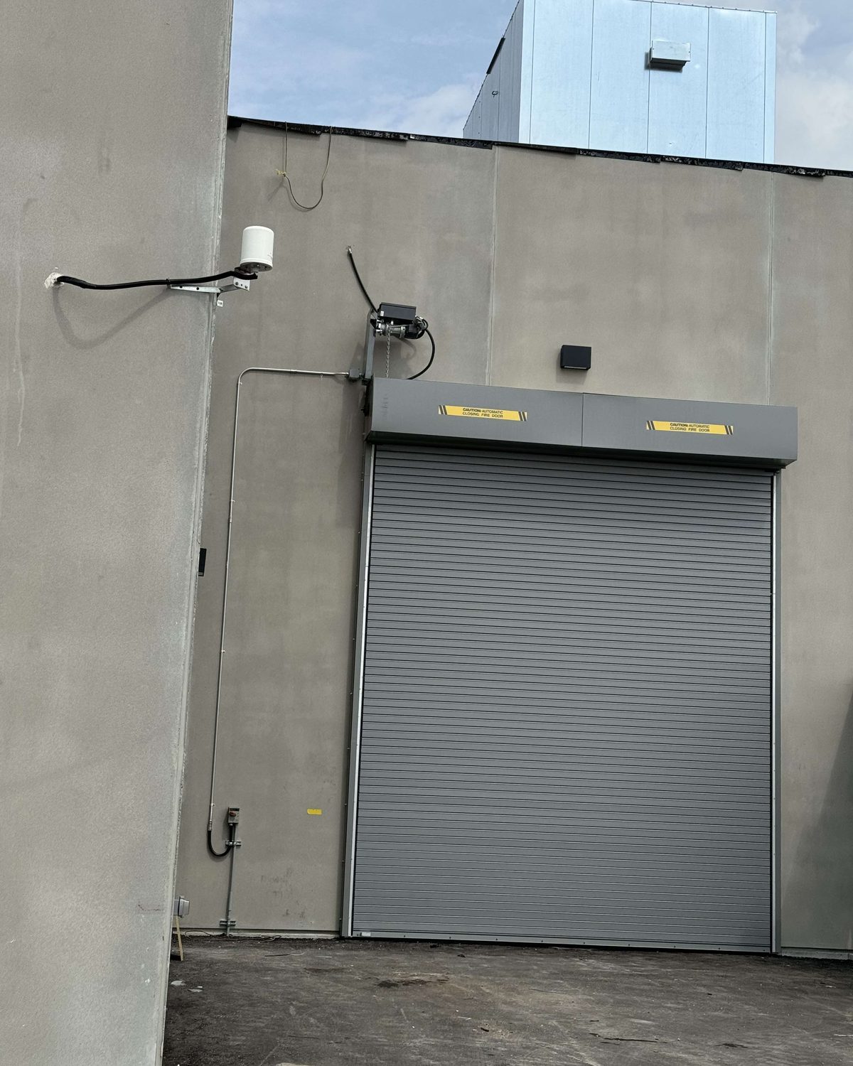 A grey rolling steel door closed in a precast concrete wall, its hood labelled with yellow caution decals reading automatic closing fire door