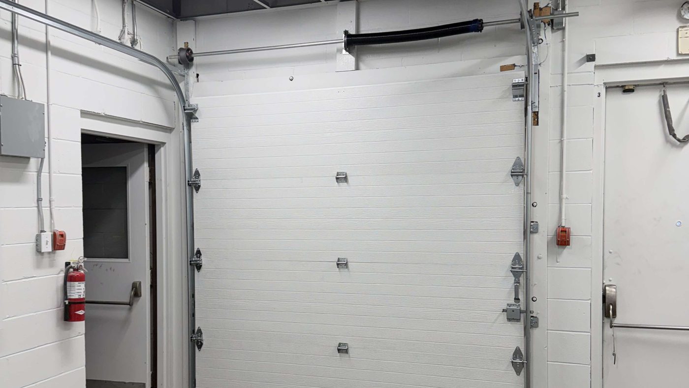 A white sectional overhead door closed in a block-walled service room, with a black torsion spring on the shaft above the door, vertical tracks at each side and a fire extinguisher on the wall