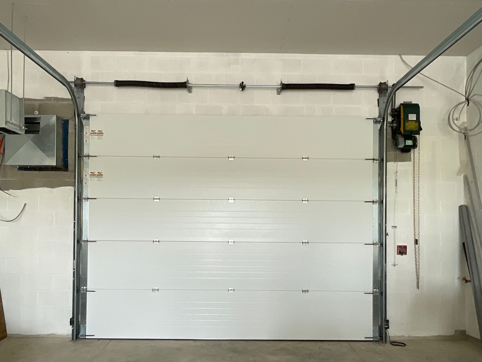 A white sectional overhead door seen from inside a new block building, with two torsion springs on a shaft above the opening, curved tracks at both sides and a wall-mounted operator with a hanging hand chain on the right