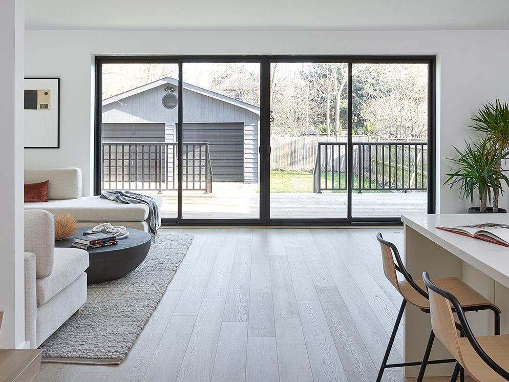 A wide black-framed sliding patio door filling the back wall of a bright open-plan living room, looking out onto a wooden deck with black railings and a grey garage beyond