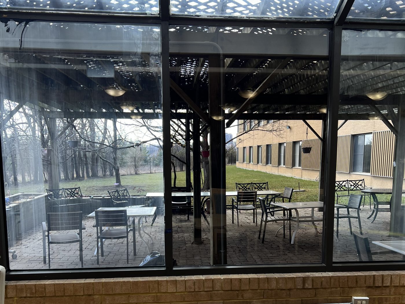 Looking out through a glazed enclosure in dark aluminum framing, its roof glazing sloping overhead, to a patio with tables, chairs and bare trees beside a commercial building