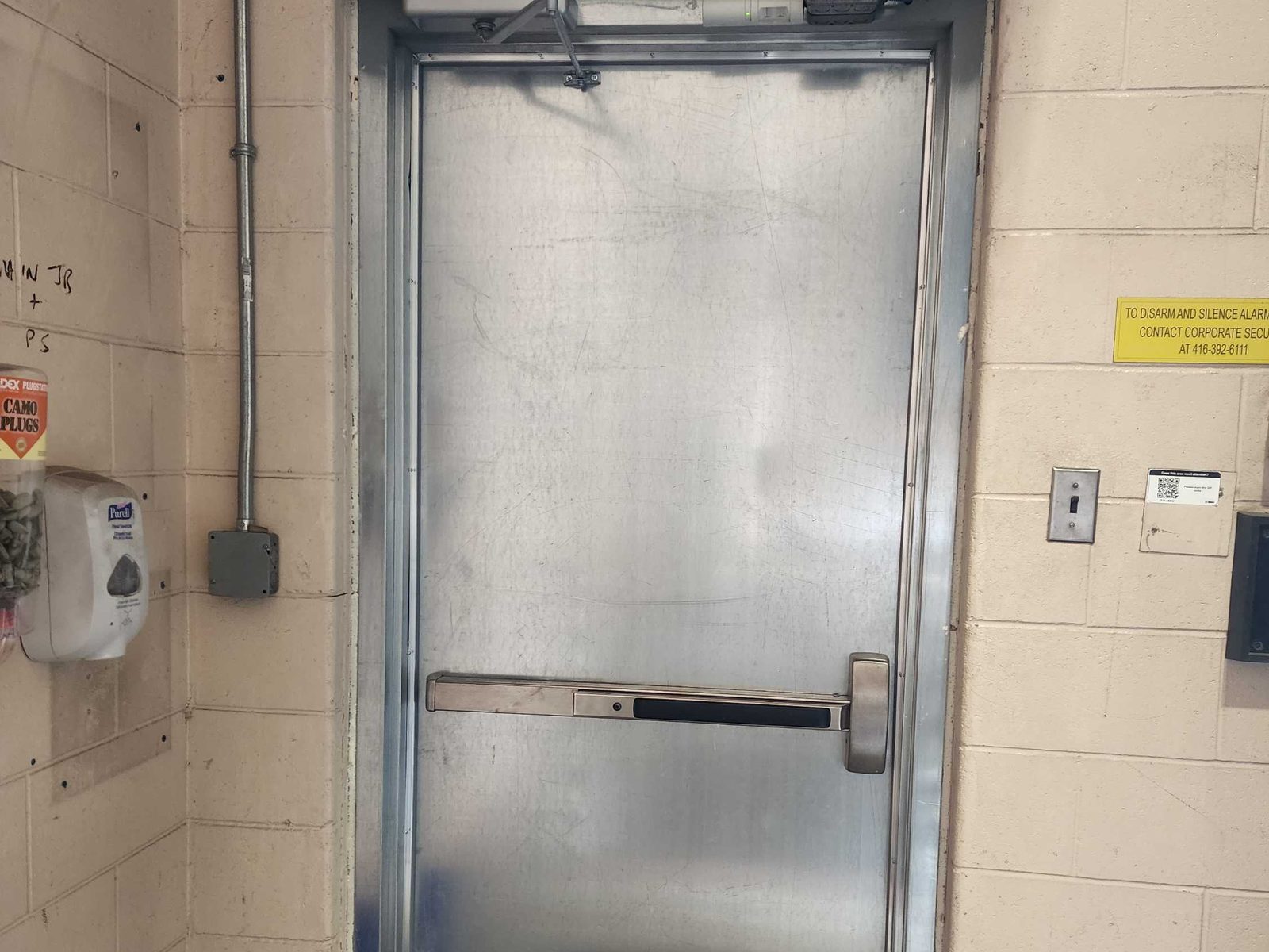 An unpainted steel door with a surface-mounted closer and a horizontal exit device, hung in a steel frame in a painted block wall, with a door-alarm notice on the wall beside it